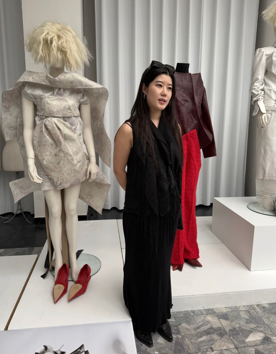 A woman in a black sleeveless outfit stands indoors between mannequins displaying fashion garments. The scene includes a mannequin with a light gray draped dress and red pointed shoes, and another with a dark red and red outfit, against a white curtain backdrop.